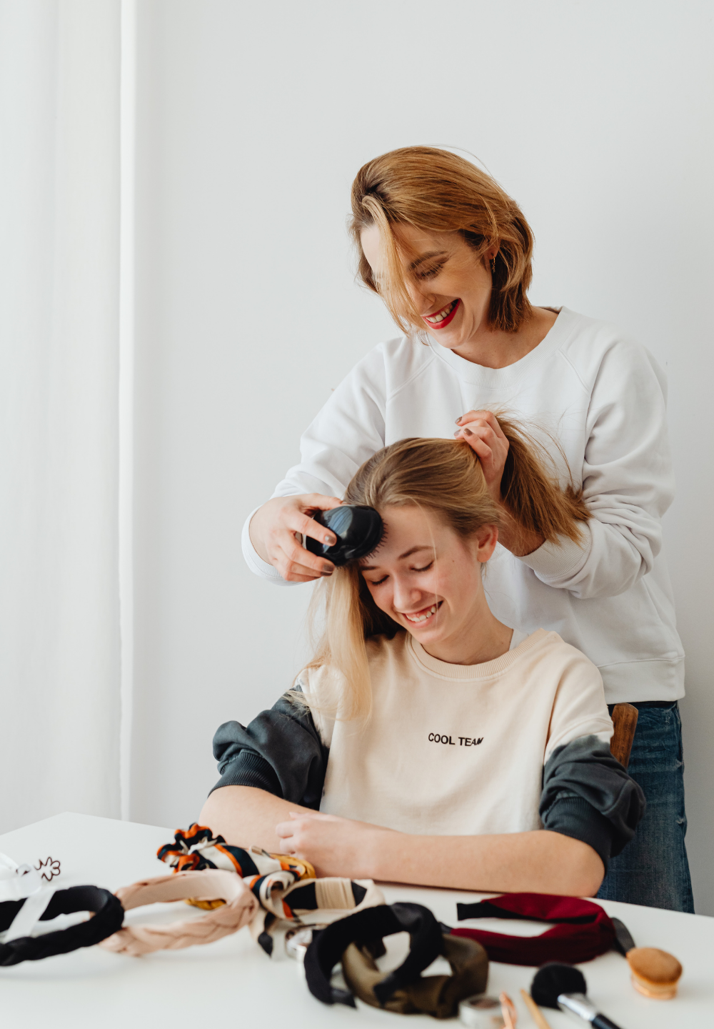 Woman brushing hair of another woman, both smiling, sitting at a table with accessories. White background.
