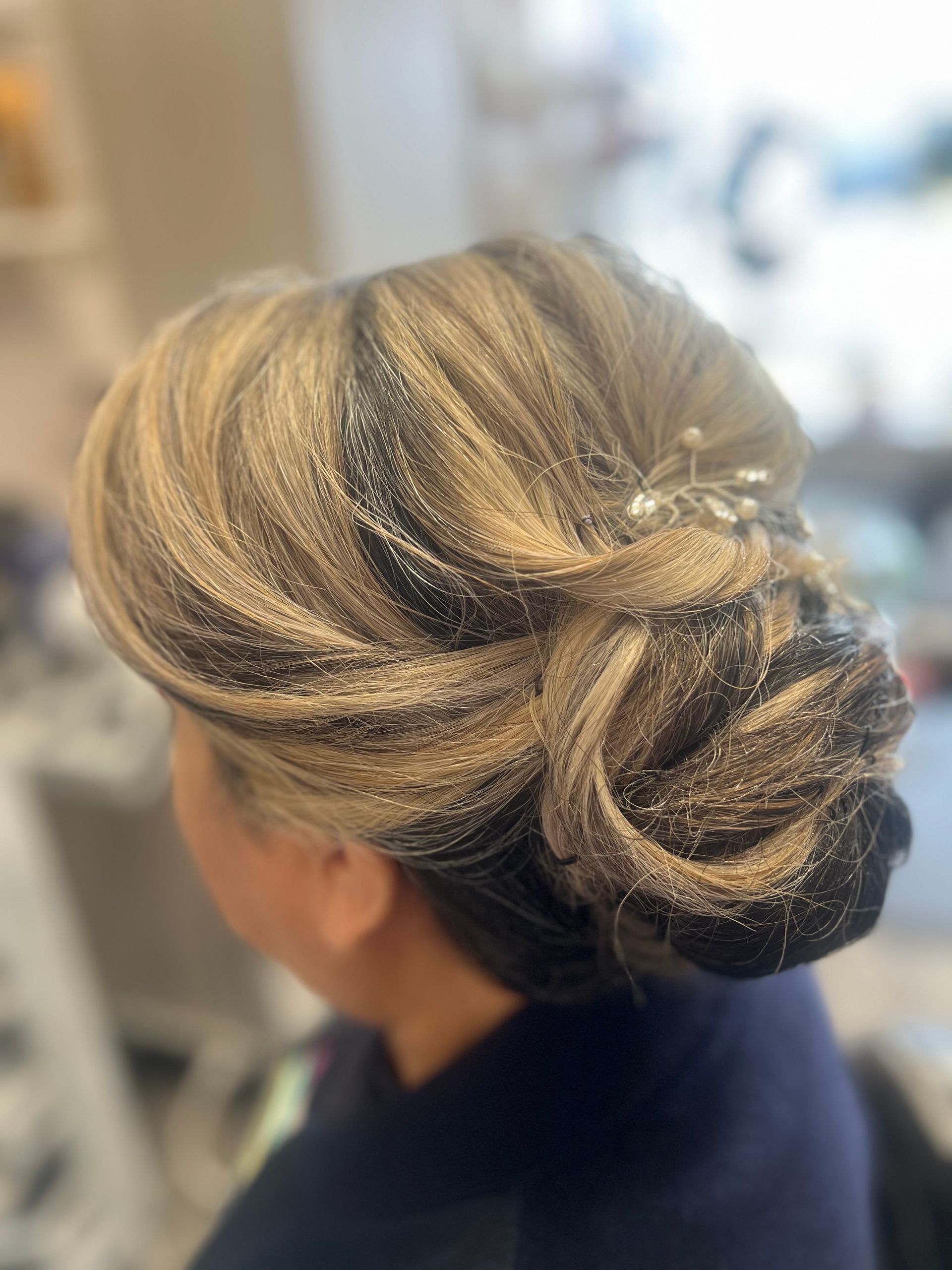 Woman with updo hairstyle being styled, wearing a silk robe. Hairdresser spraying hair near a brick wall.