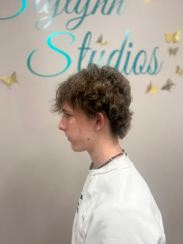 A person with a mullet hairstyle stands sideways against a wall, facing left.