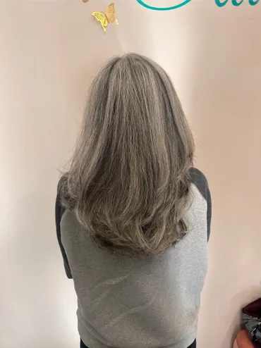 Woman with long, gray hair, wearing a gray and black shirt, standing with back to camera. Pink wall background.