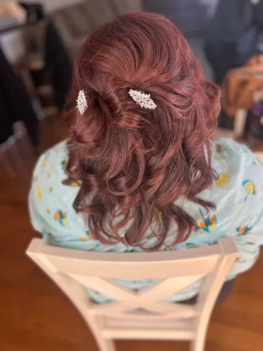 Woman with auburn hair styled in a half-updo, held with two decorative hairpins, seated in a chair.