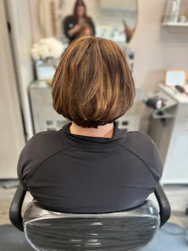 Woman with brown hair in a salon chair, back view, highlights, short bob, black shirt, salon setting.