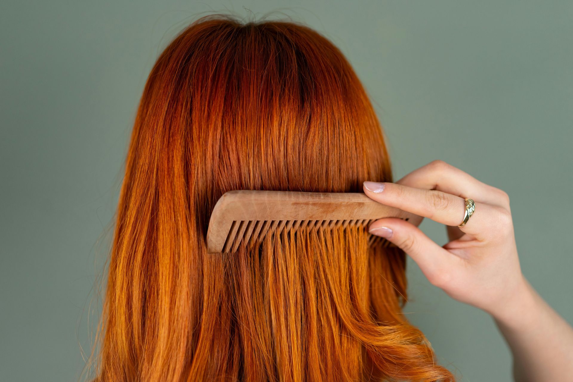 Person combing vibrant red hair with a wooden comb against a muted green background.