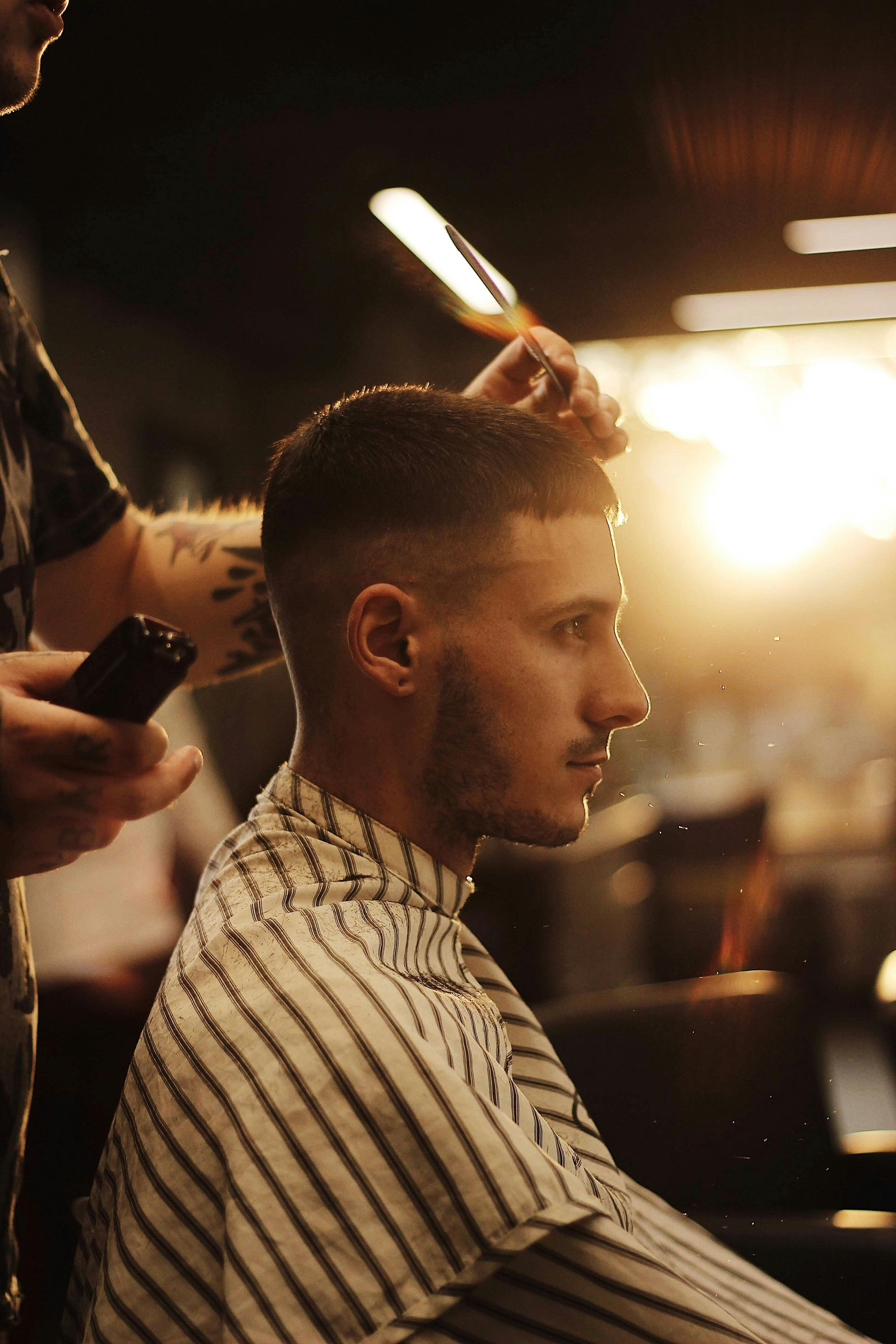 Man getting a haircut in barbershop, wearing striped cape. Barber using straight razor. Backlit setting.