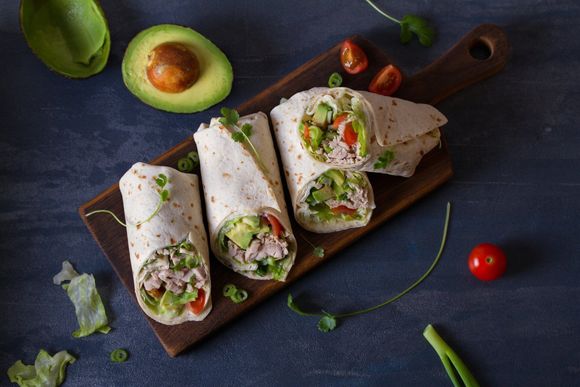 three tortilla wraps with avocado , tomatoes and lettuce on a wooden cutting board .
