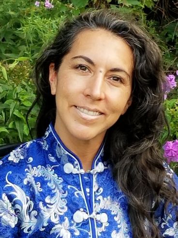 A woman in a blue floral shirt is smiling in front of flowers.