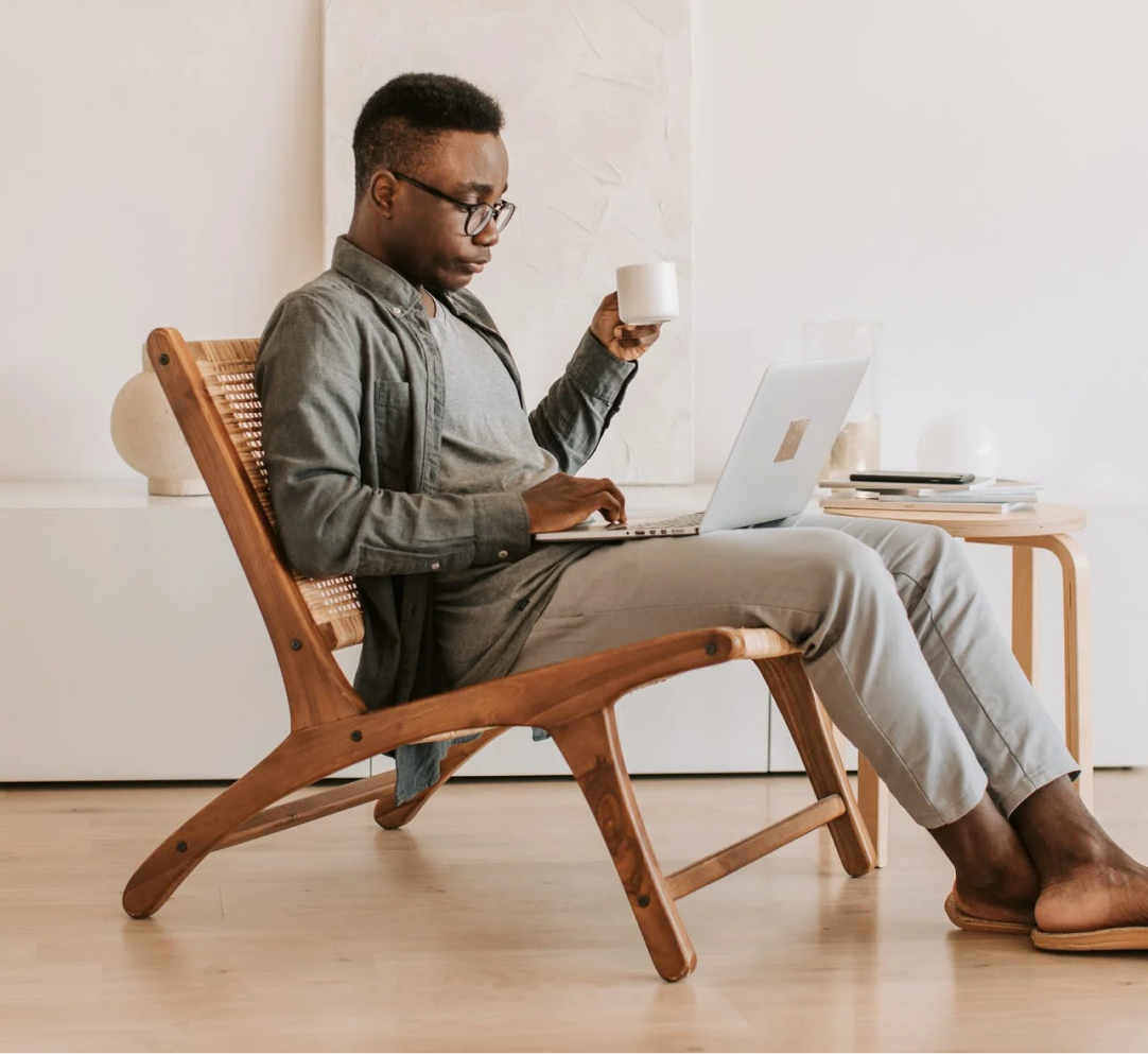 A man is sitting in a chair using a laptop computer while holding a cup of coffee.