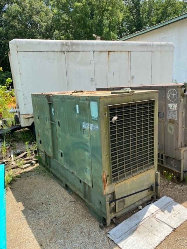 A large green generator is sitting in a dirt lot next to a white trailer.