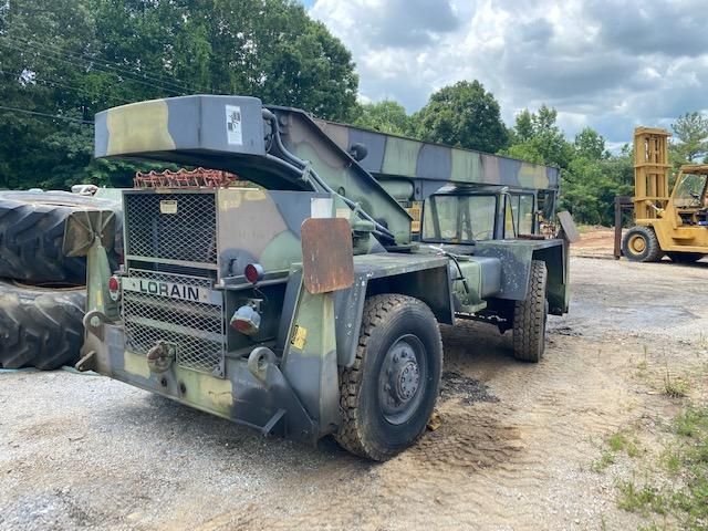 A military truck is parked in a dirt lot next to a forklift.