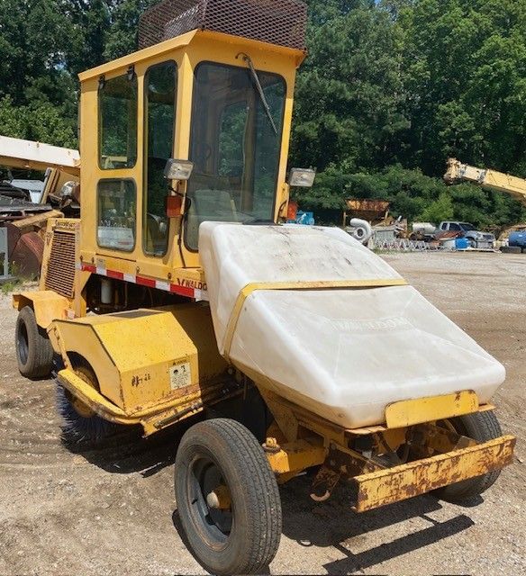 A yellow tractor with a white tank on the back is parked in a dirt lot