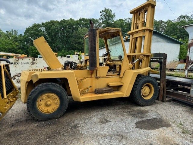 A yellow forklift with its hood up is parked in a gravel lot