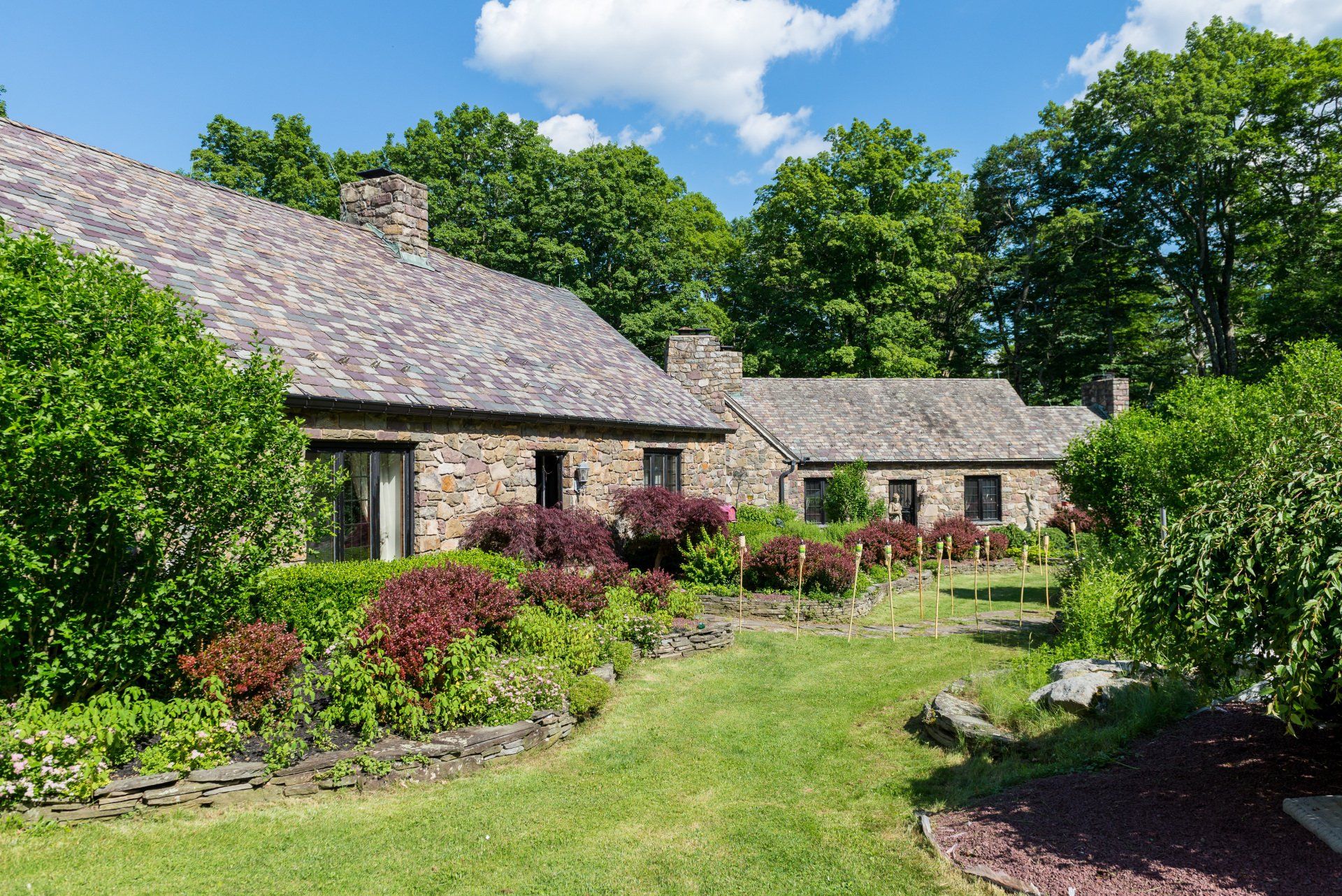 A large stone house with a slate roof is surrounded by trees and bushes.