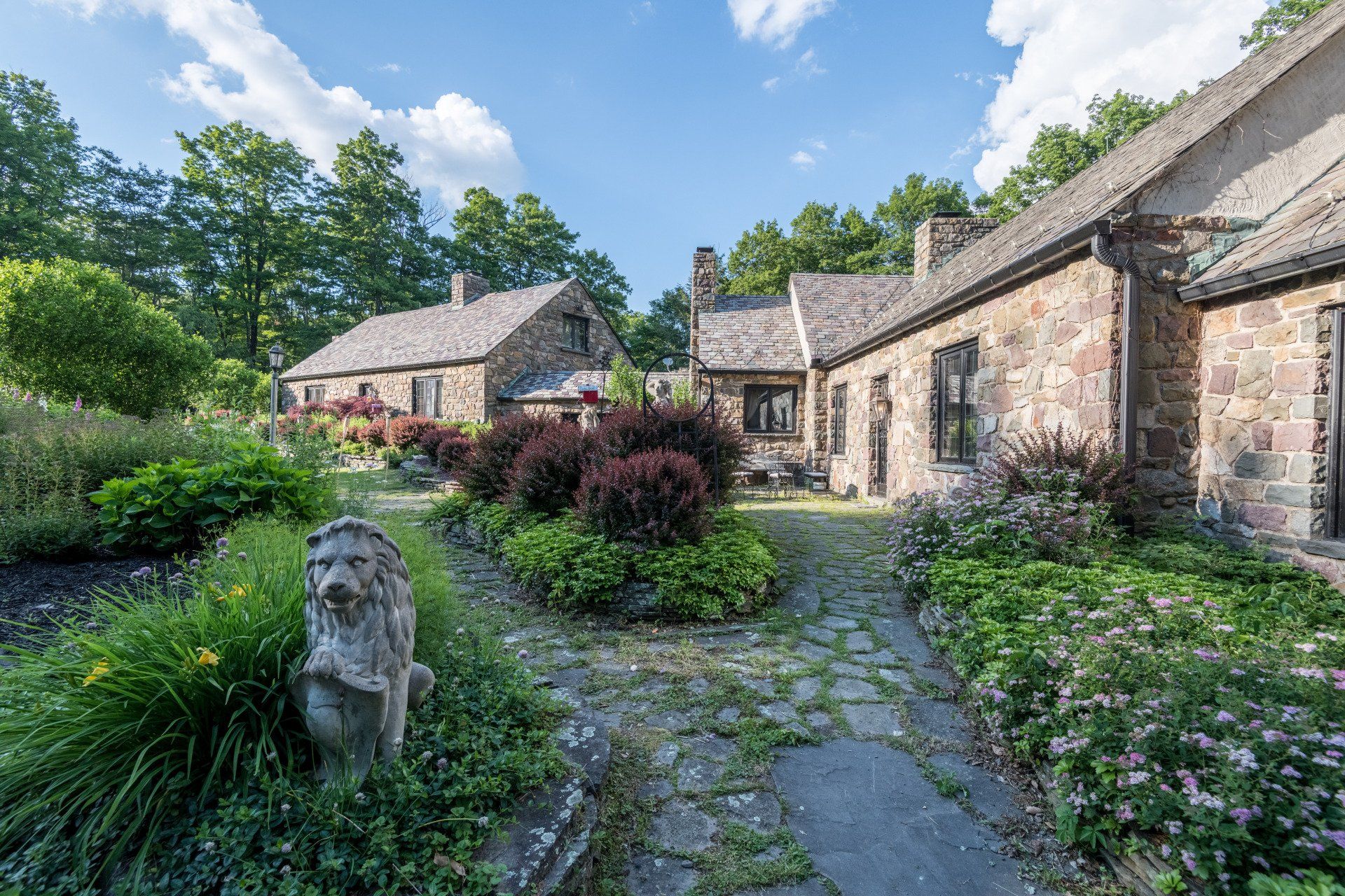 A stone statue of a lion is in the middle of a garden in front of a stone house.