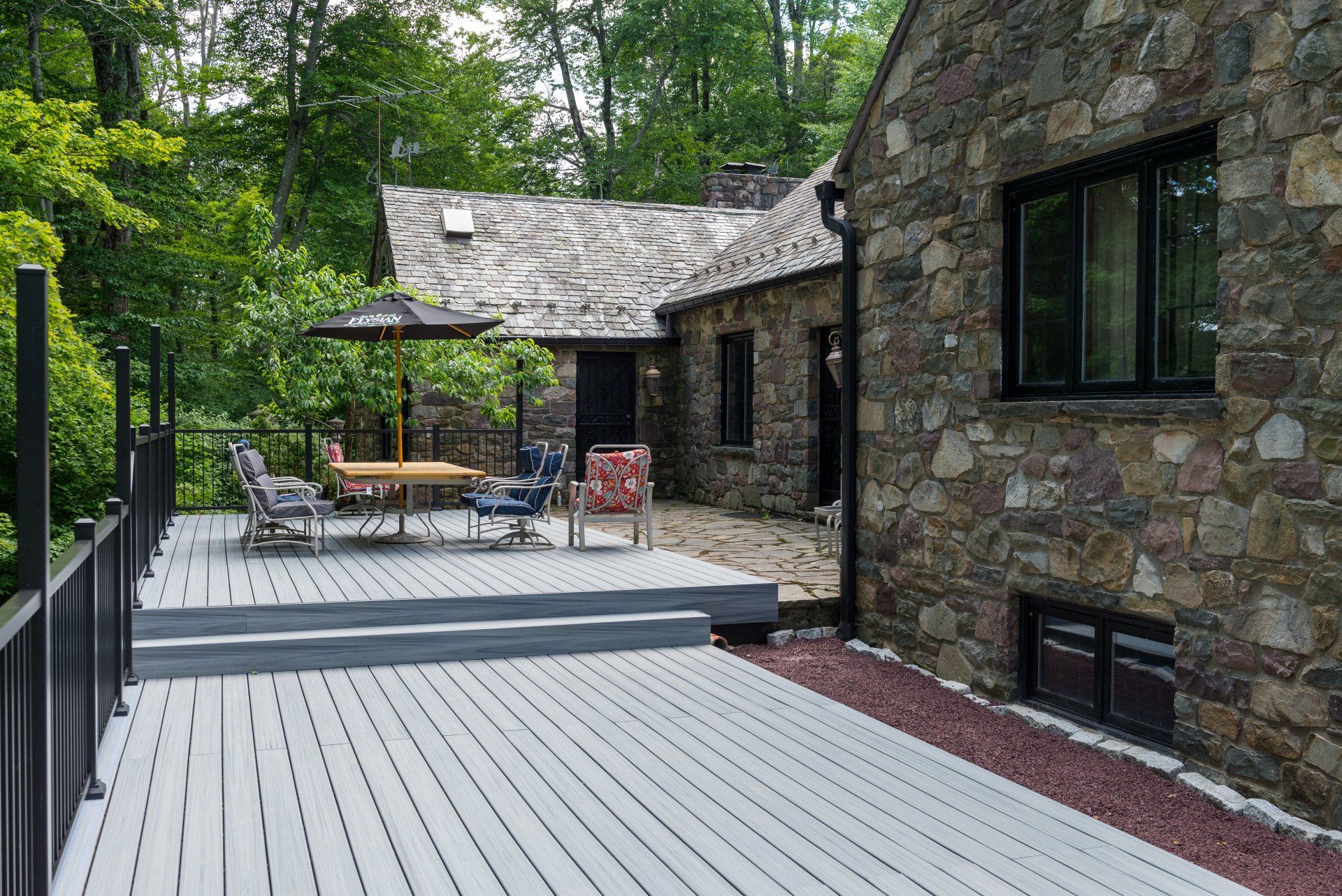 There is a large deck in front of a stone house with a table and chairs on it.