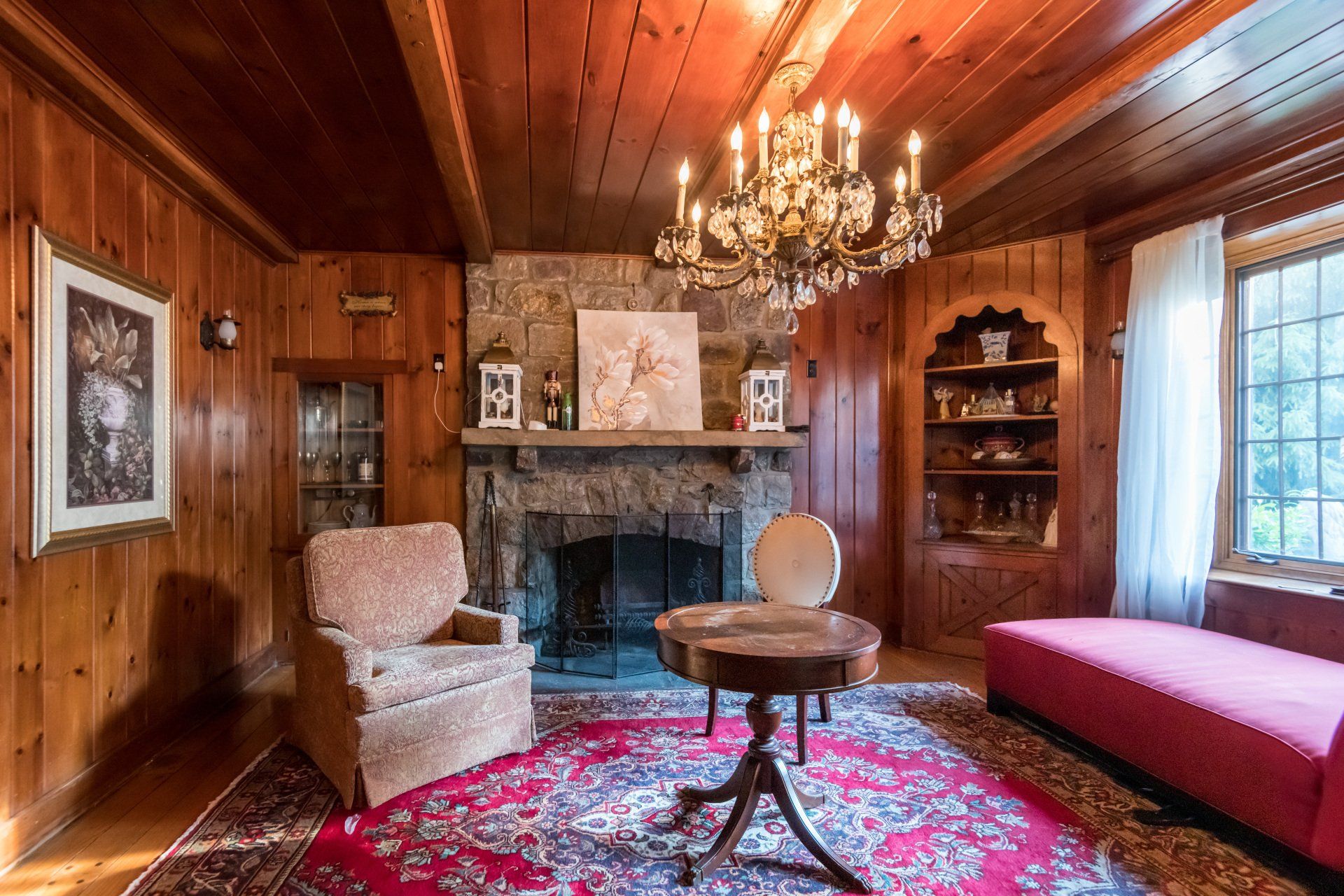A living room with wood paneling , a fireplace , chairs , a table and a chandelier.