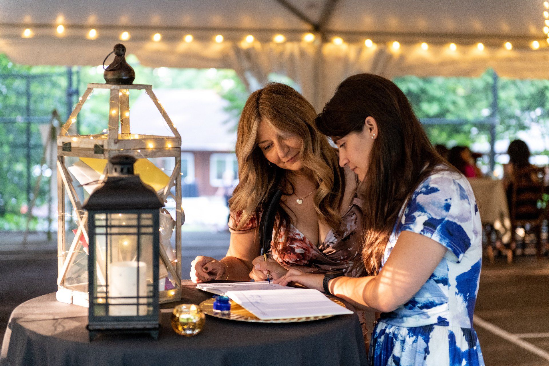 Two women are signing a document at a table under a tent.