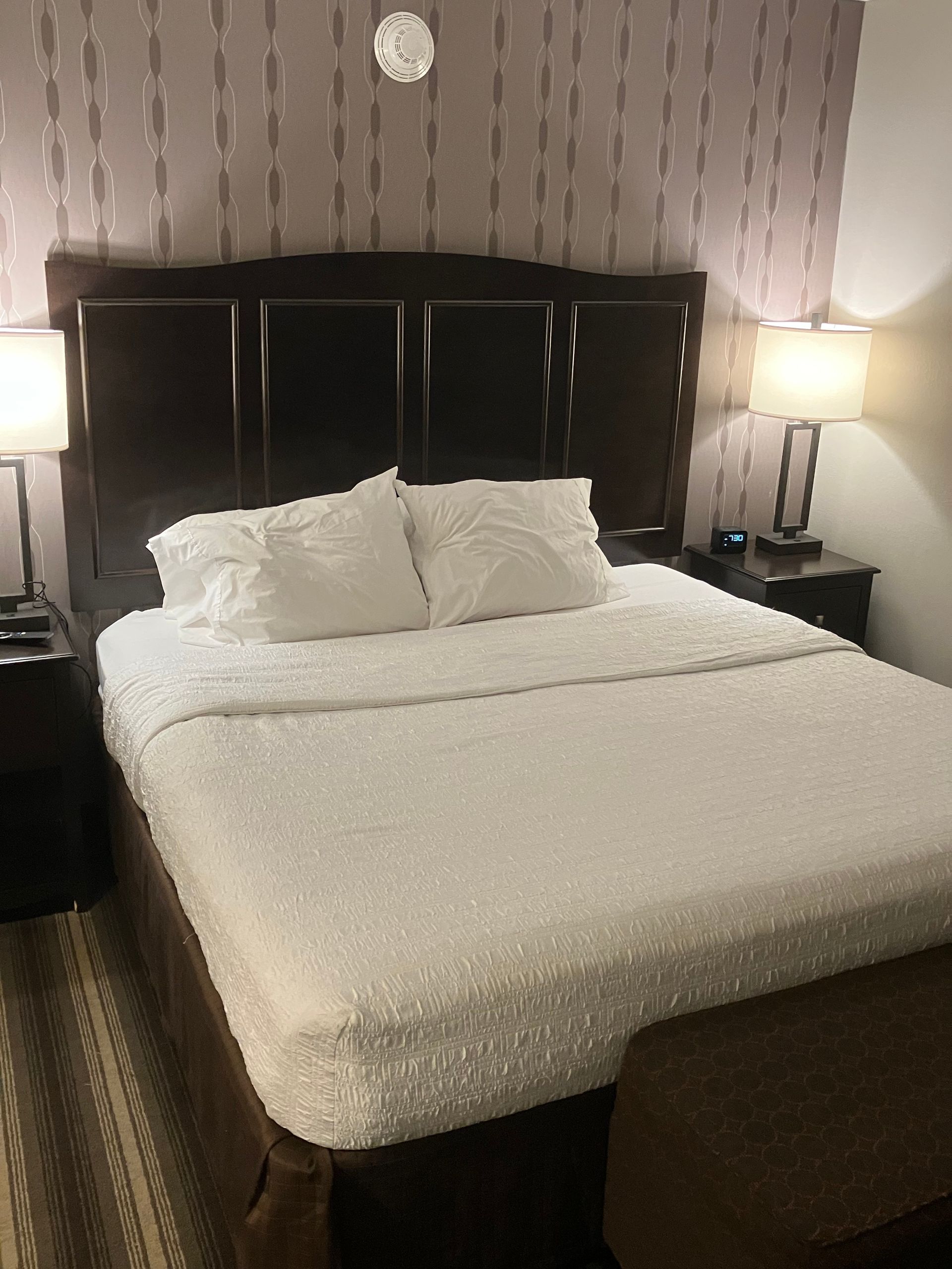 Hotel room with a made bed, bedside tables with lamps, and patterned wallpaper.