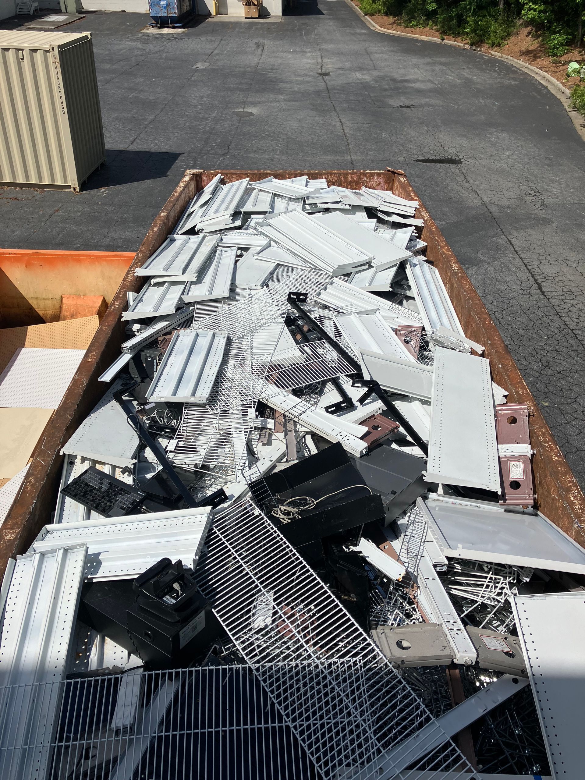 Scrap metal and computer parts fill a large dumpster outdoors on asphalt.