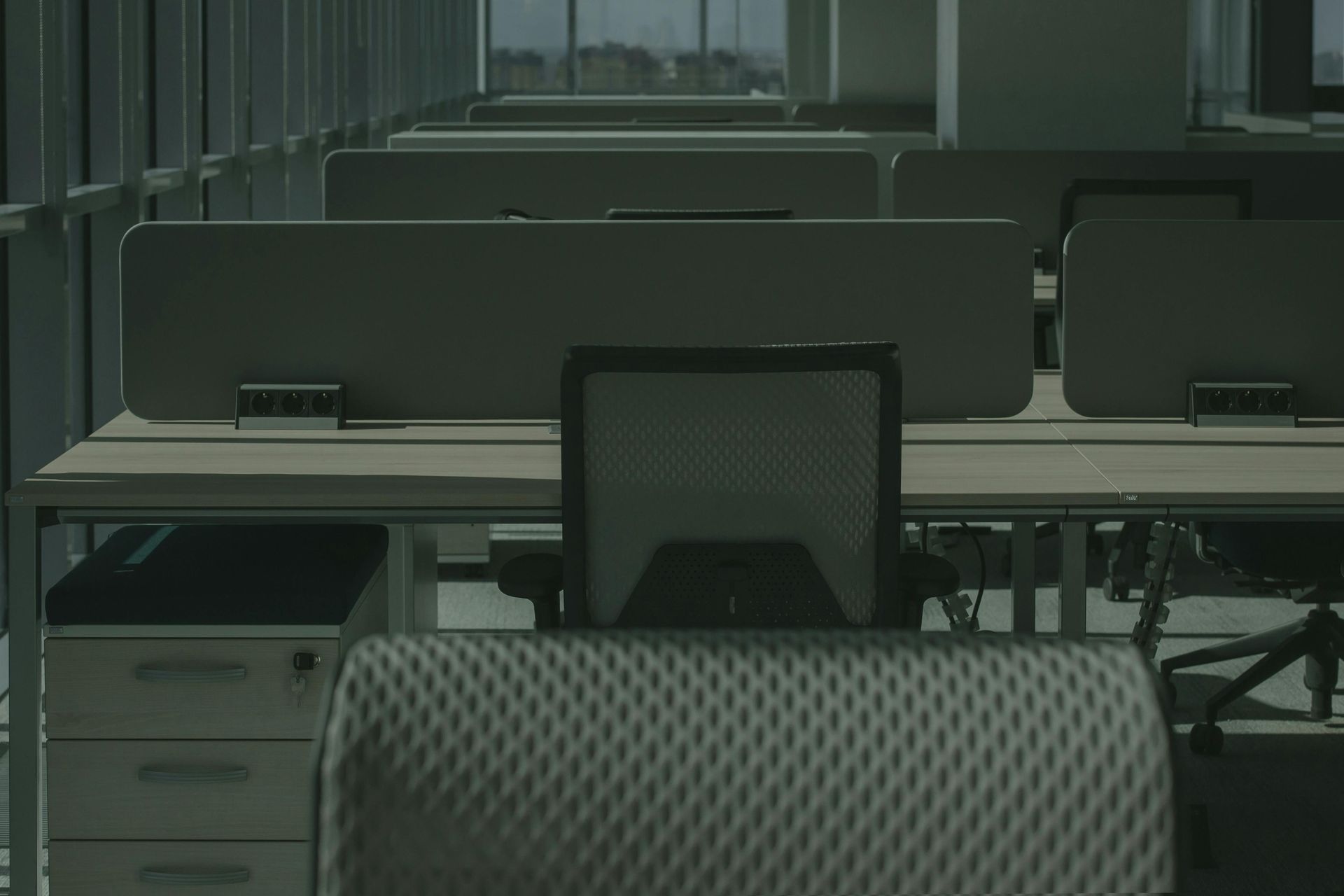Office interior: wooden desk and cabinet, chairs, patterned carpet, large window, brown walls, and ceiling lights.