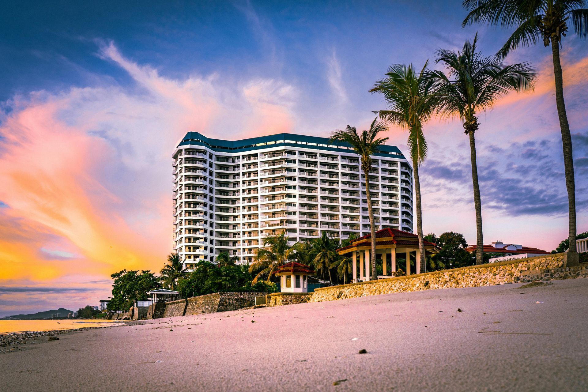 Beachfront resort at sunset; palm trees, white building, colorful sky.