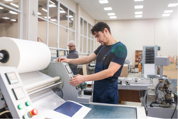 Man operating a printing machine in a workshop, another person in the background.