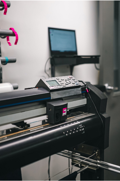 A large format printer with a control panel and connected to a laptop on a small shelf.
