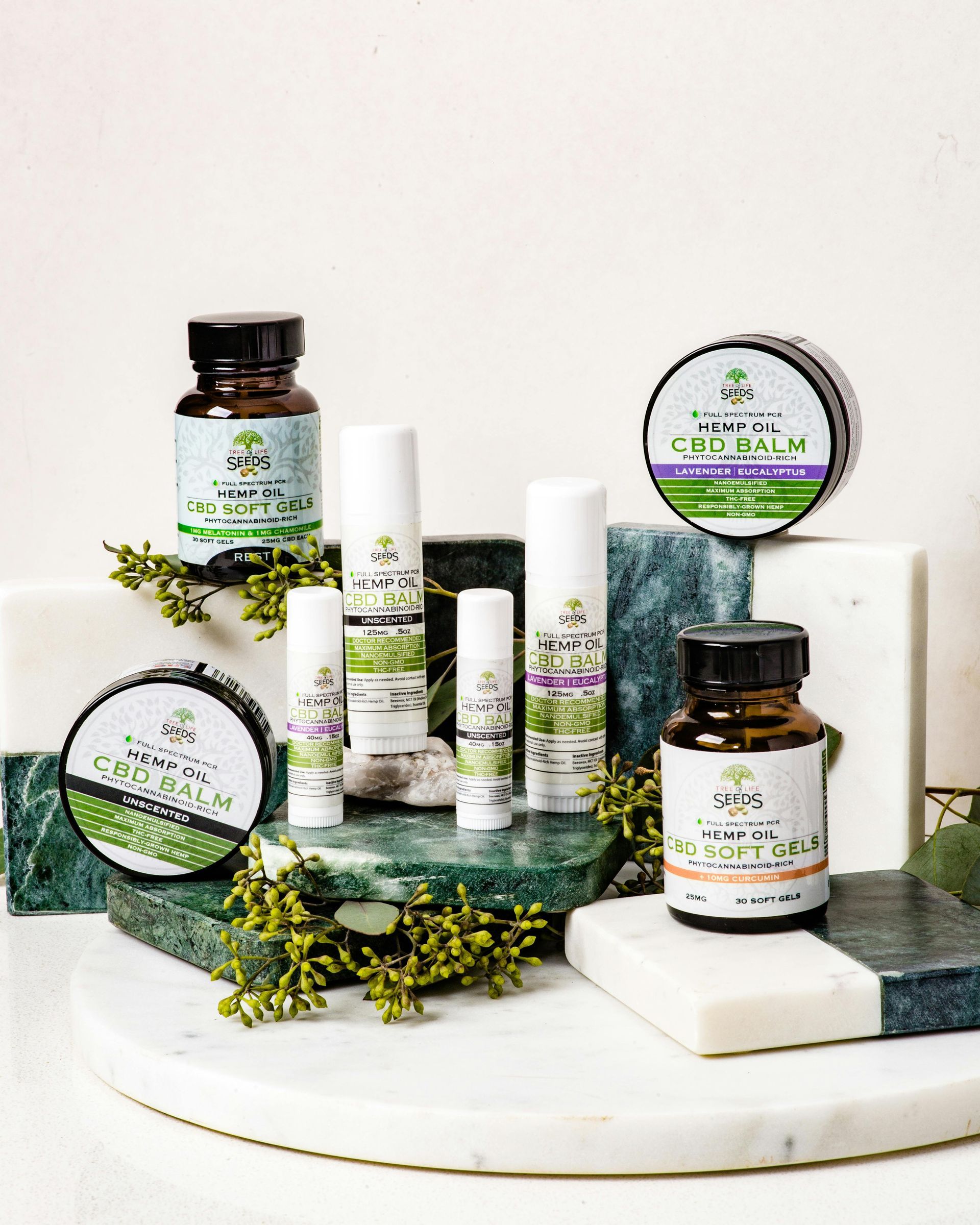 A collection of wellness products, including jars and balms, arranged on white and green marble blocks with green foliage.
