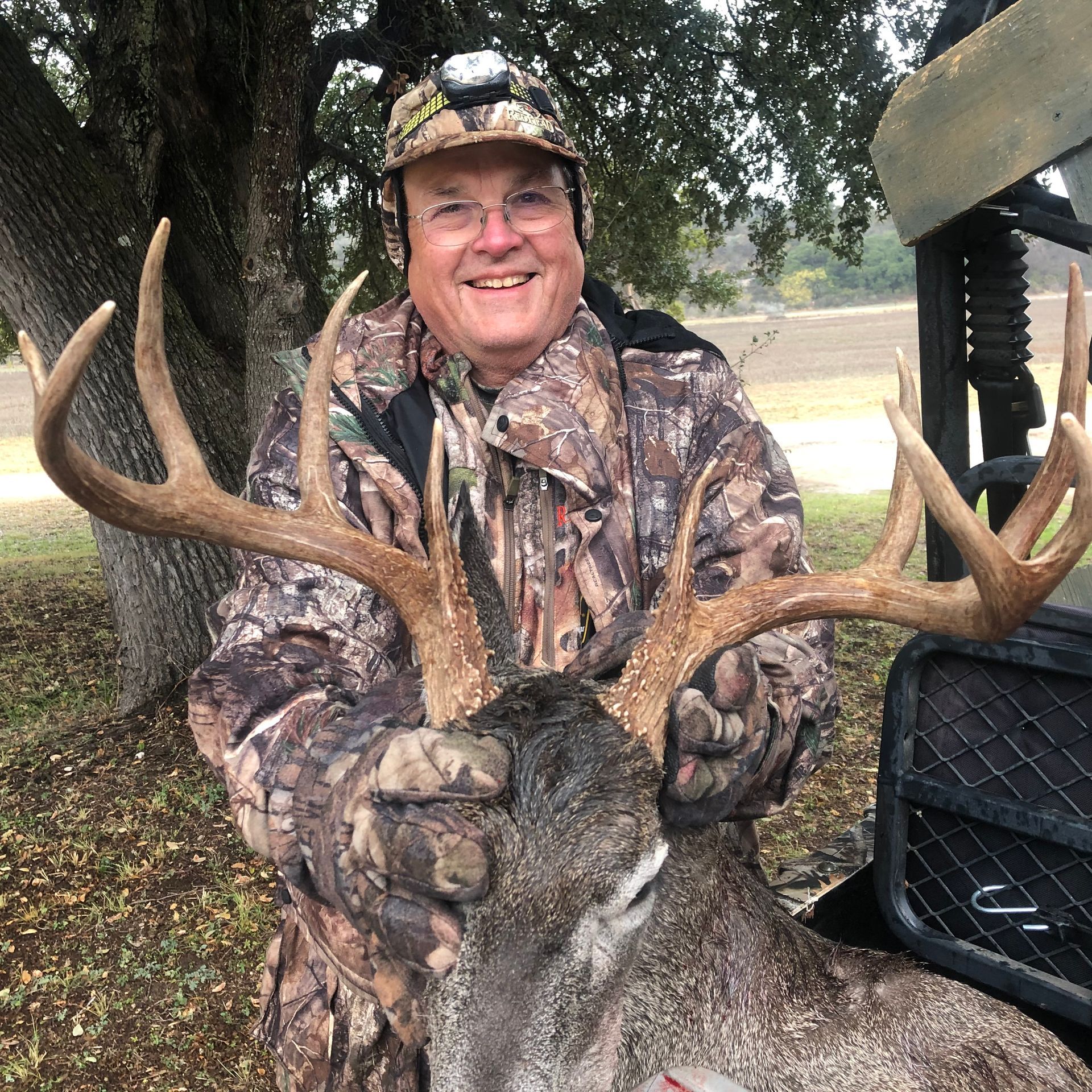 Man in camouflage holding a large buck with antlers; outdoors, smiling.