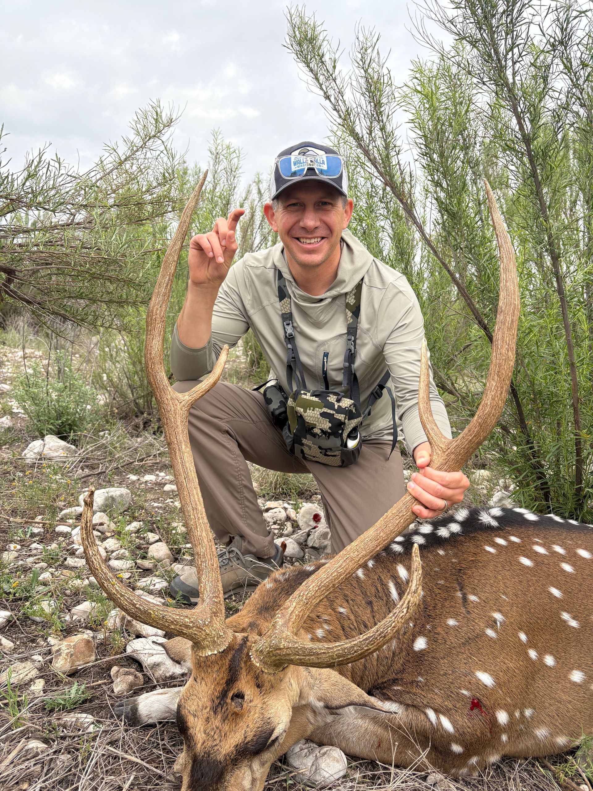 Man kneeling beside a spotted deer, holding its antlers. He wears camouflage and a hat, smiling. Outdoors.
