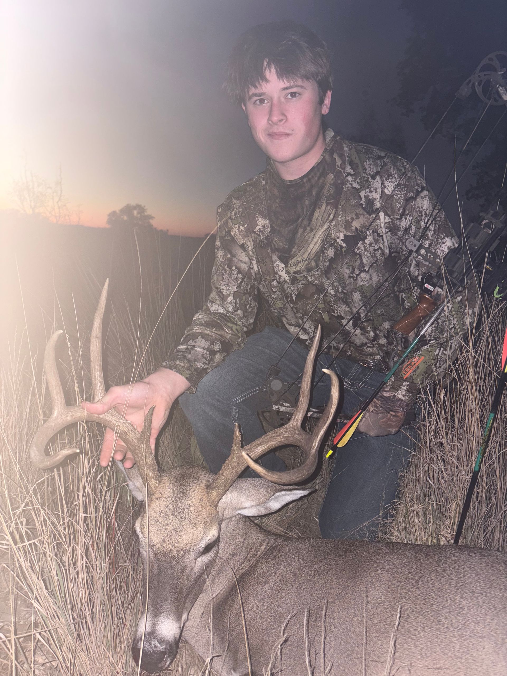 Teenager in camouflage kneels beside a harvested buck with large antlers, holding a bow and arrow.