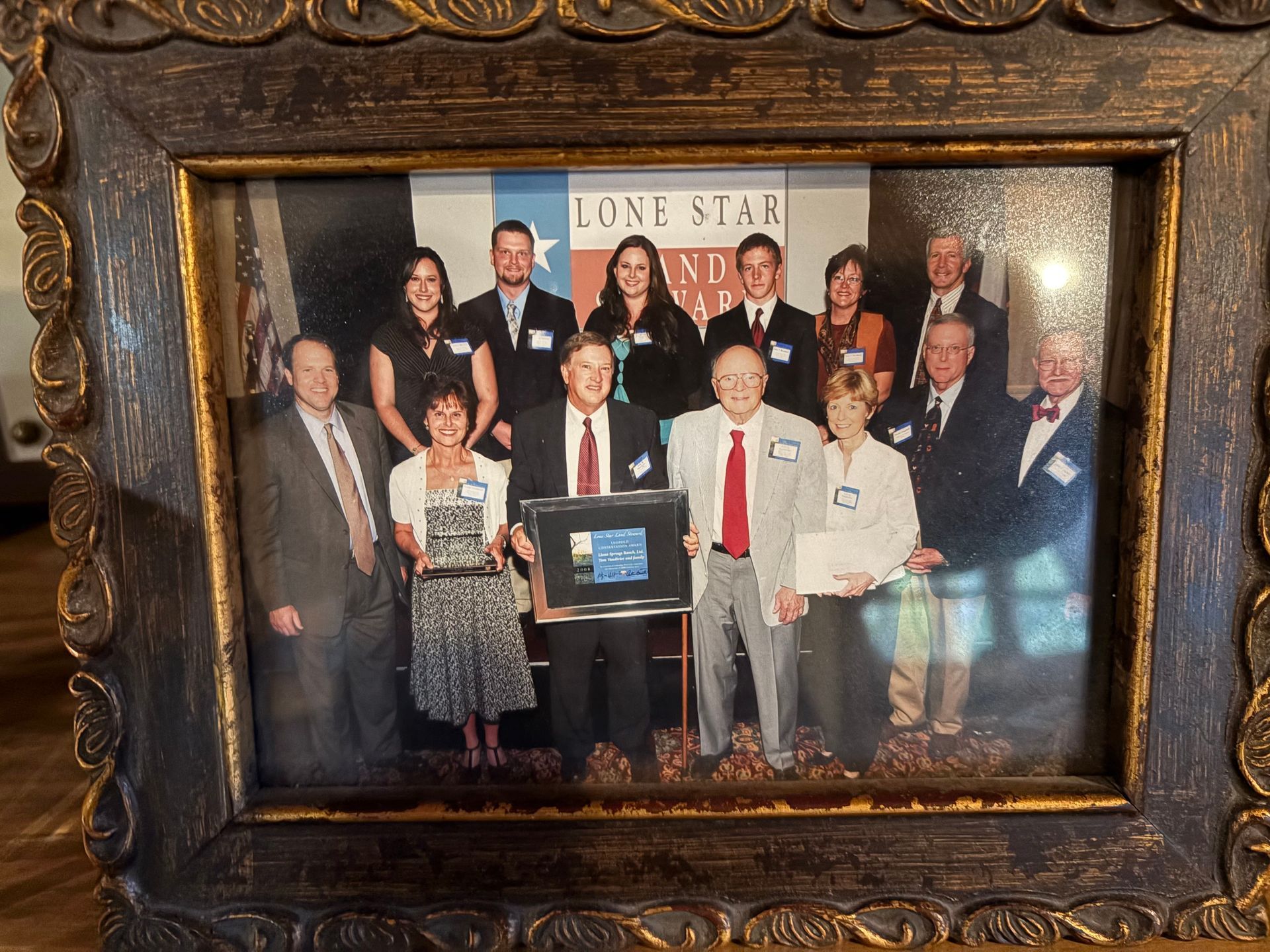 Group photo with framed award, Lone Star Land Bank banner backdrop. People in formal attire, smiling.