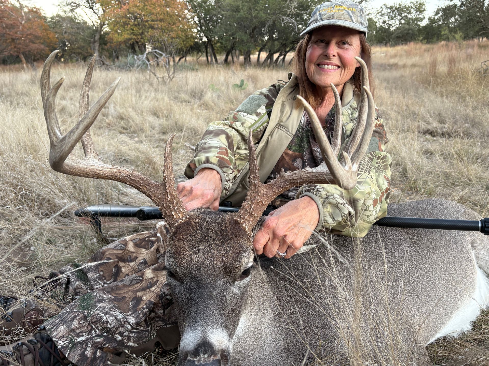 Woman in camouflage with harvested buck, smiling, in a field, holding antlers.