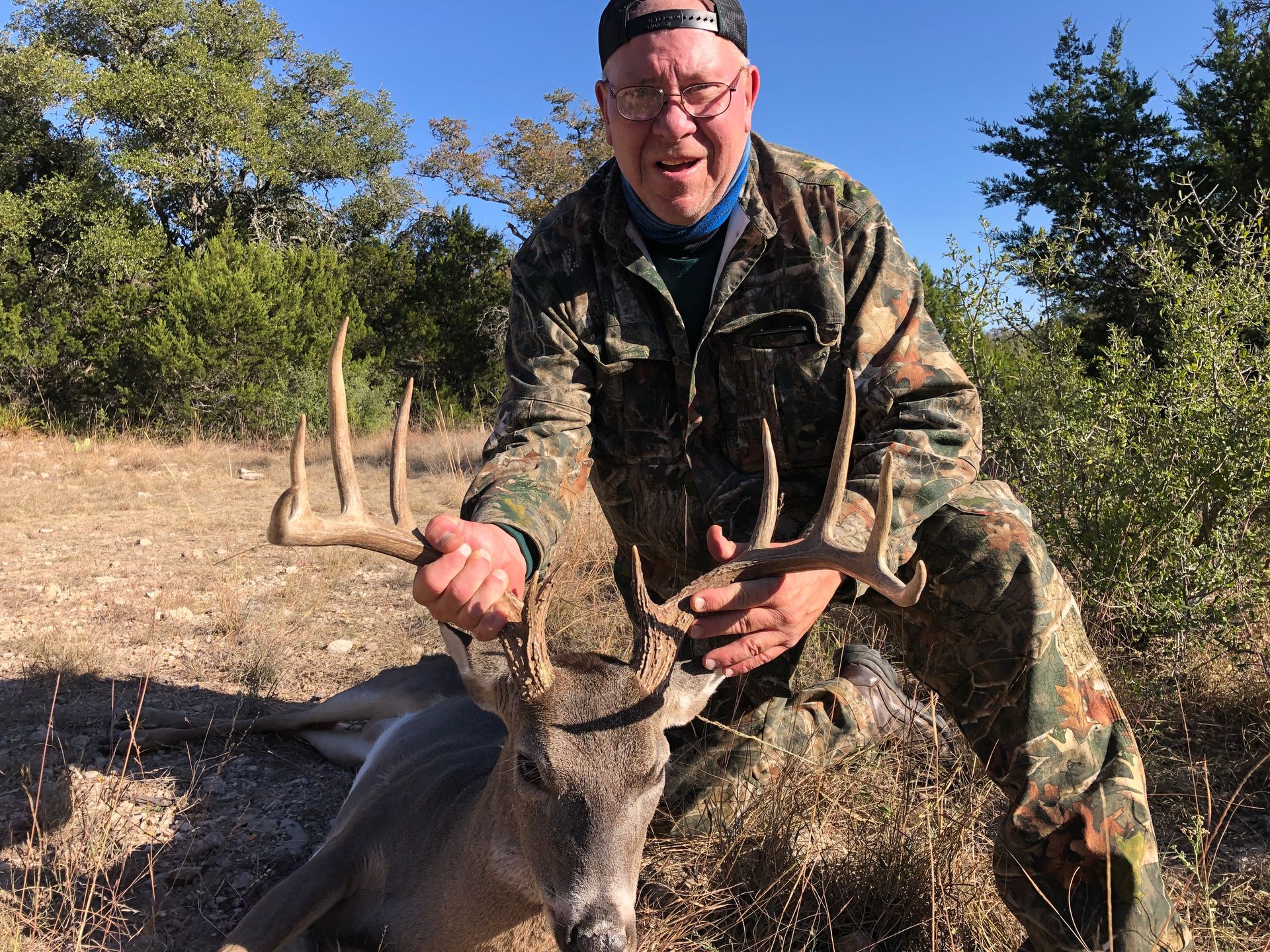 Man in camouflage holds up antlers next to a harvested deer in a wooded area.