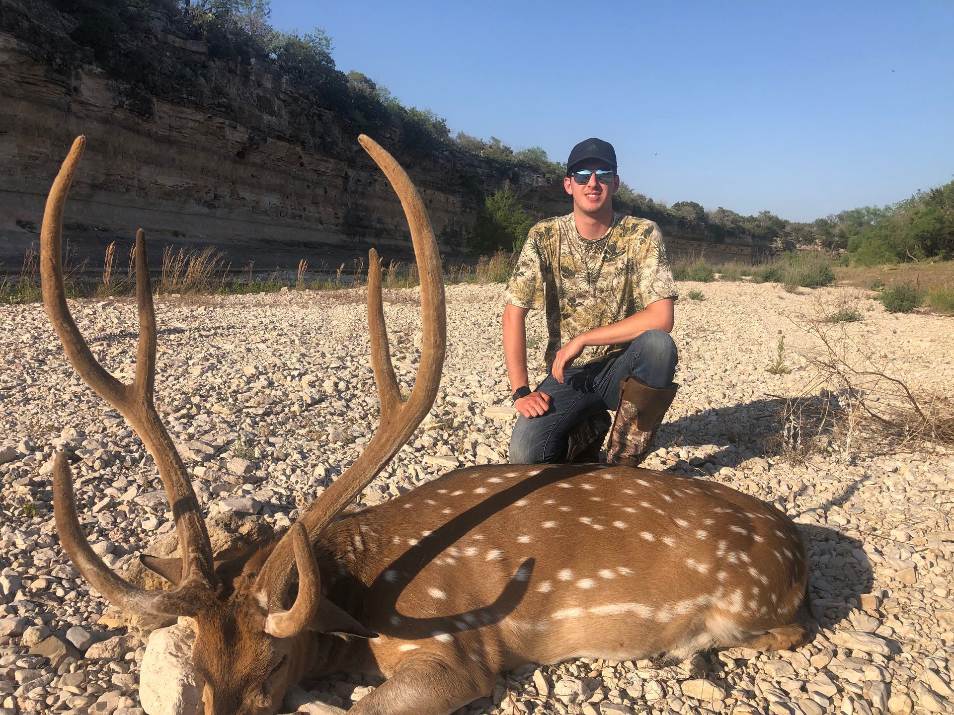 Hunter kneels beside a spotted deer with large antlers in a rocky riverbed.