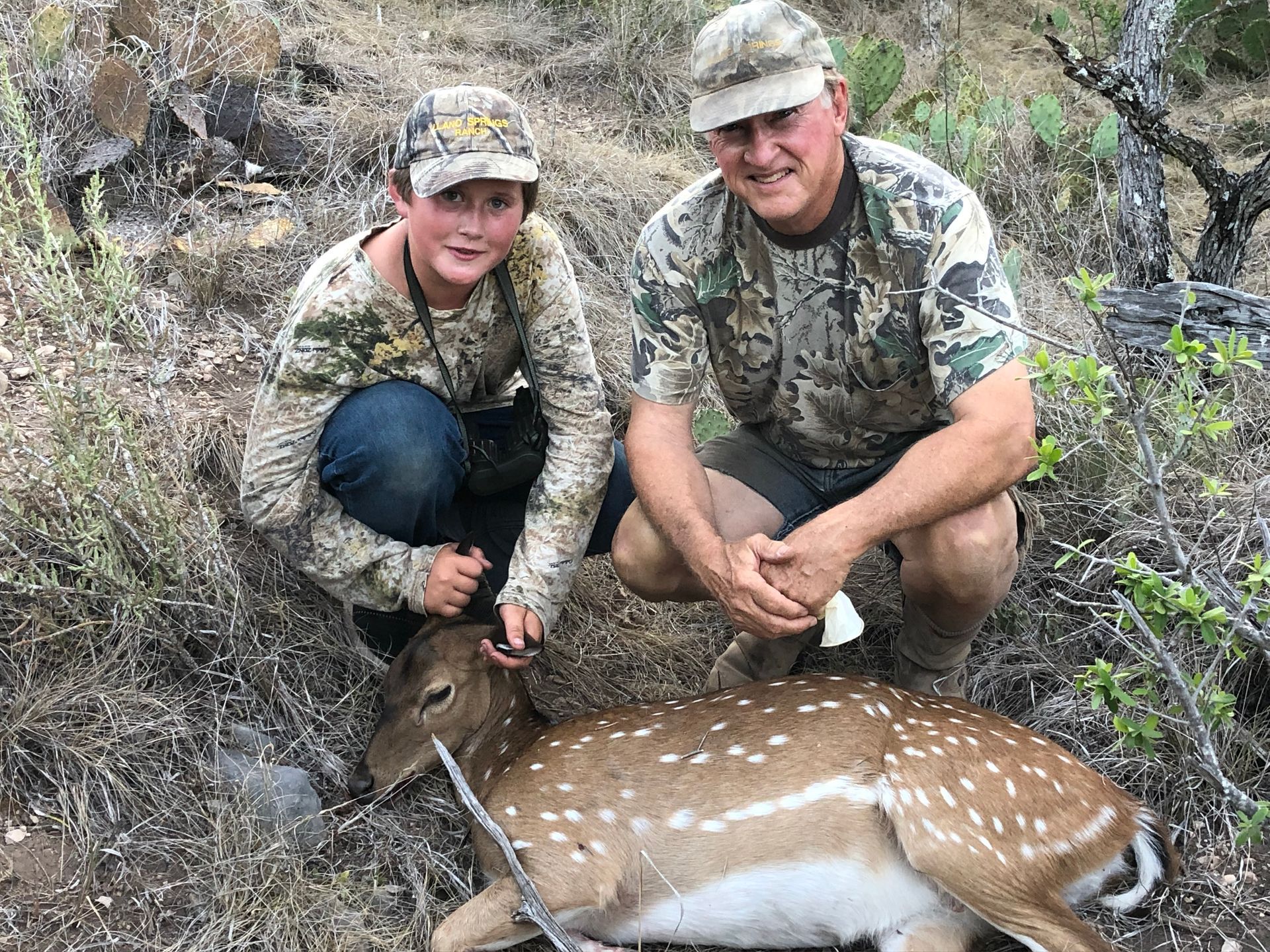 A boy and man in camouflage pose with a spotted deer in a natural setting.