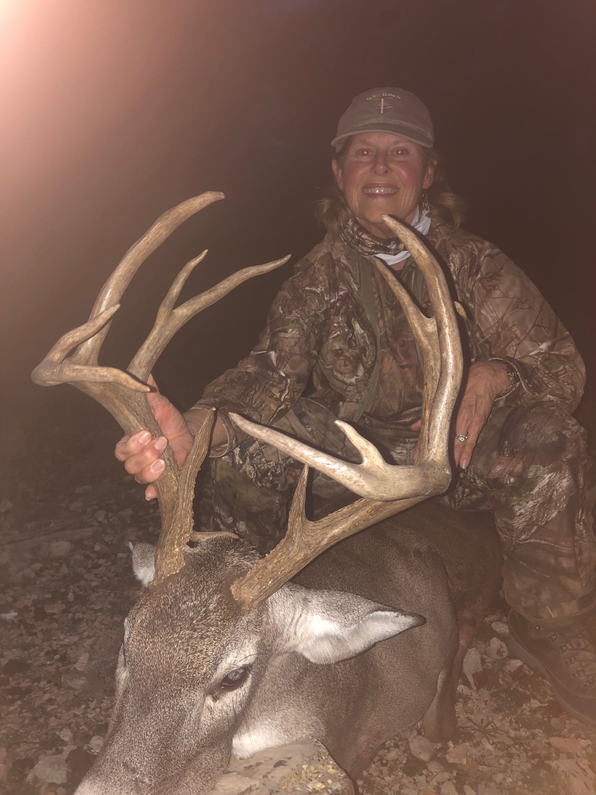 Woman in camo kneels beside a buck with large antlers, smiling outdoors at night.