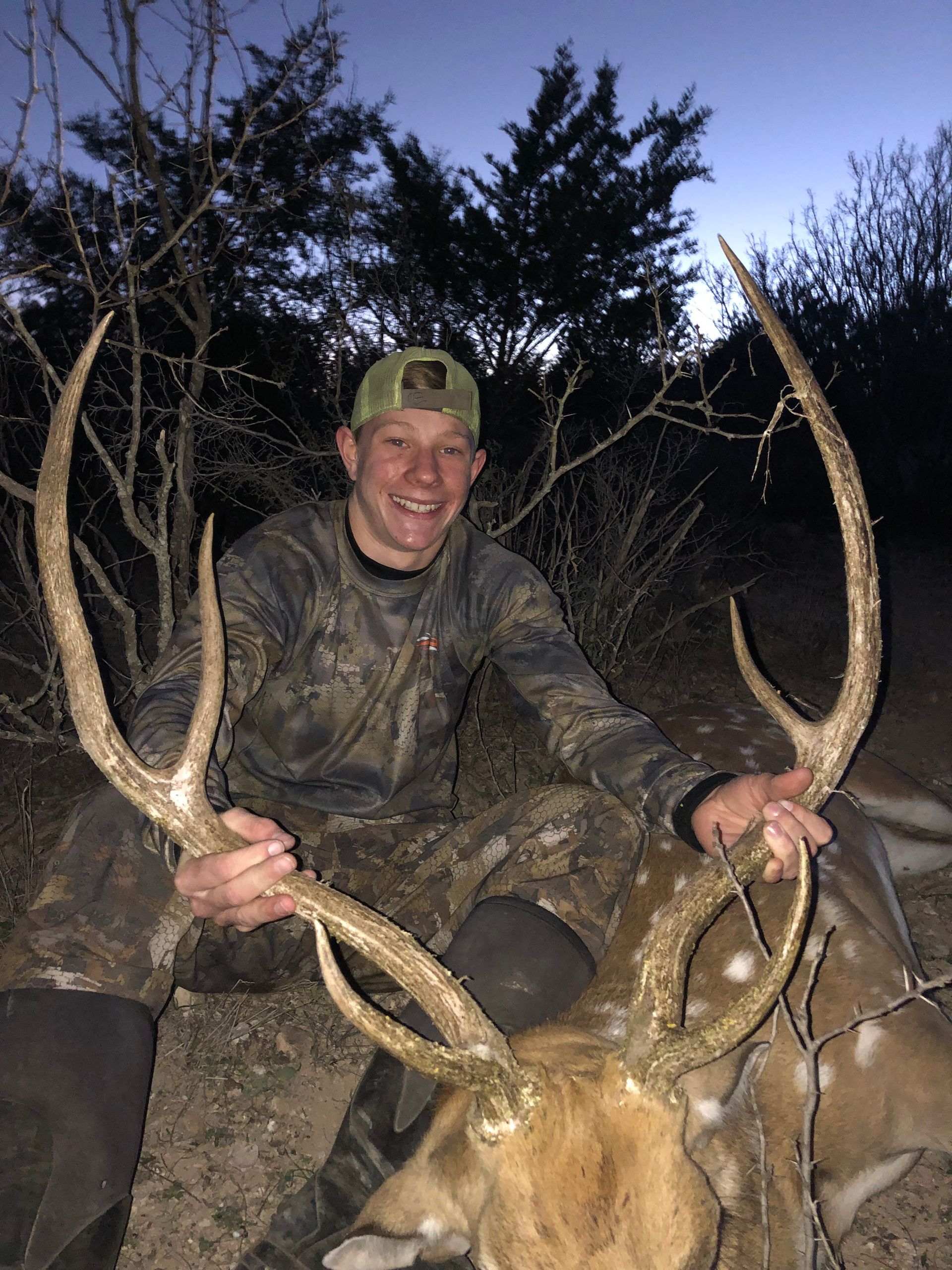 Hunter kneels, holding large antlers of a fallen deer; outdoors at dusk. He smiles, wearing camouflage clothing and a cap.