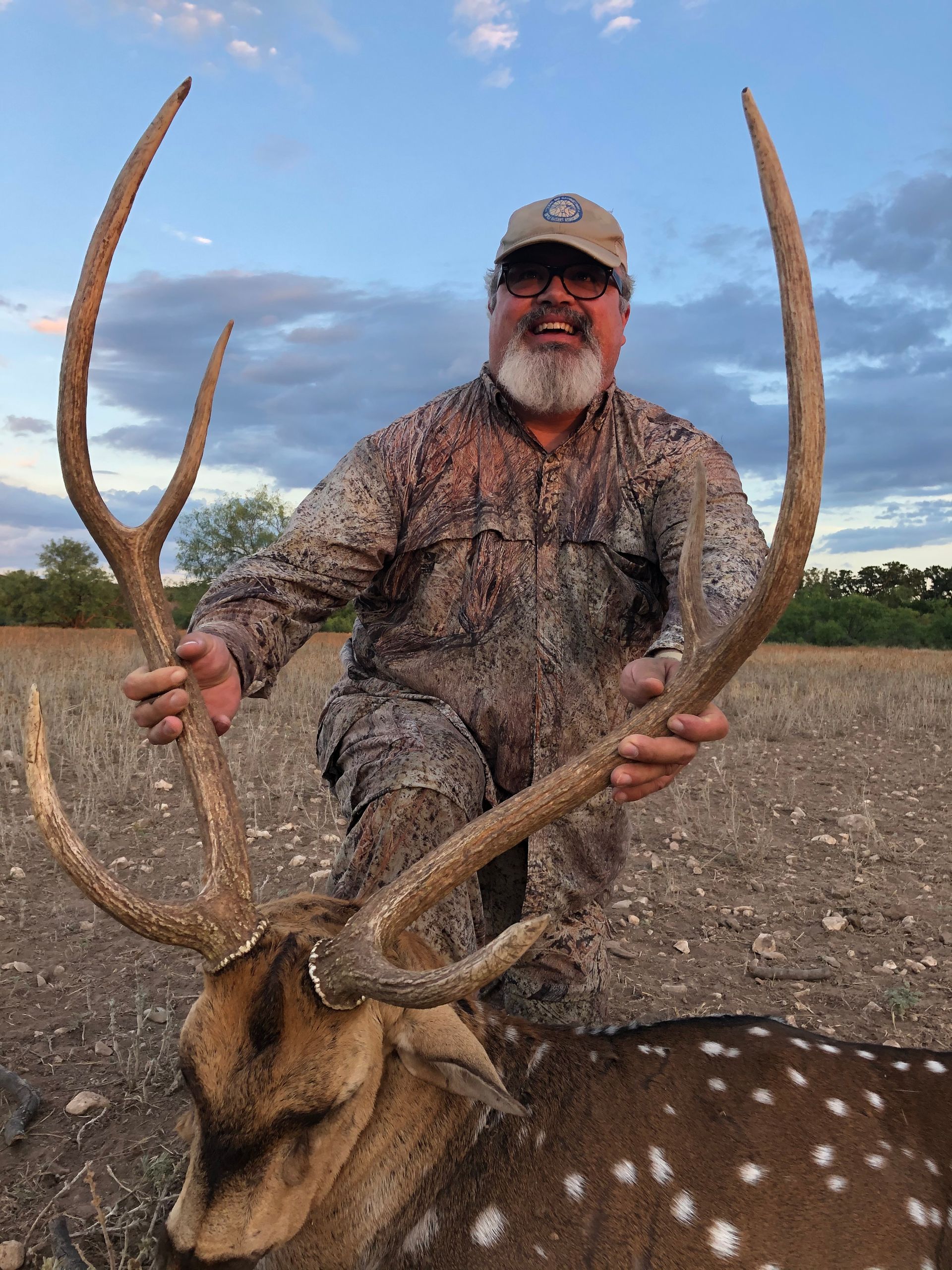 Man in camouflage holding large antlered deer, smiling outdoors.