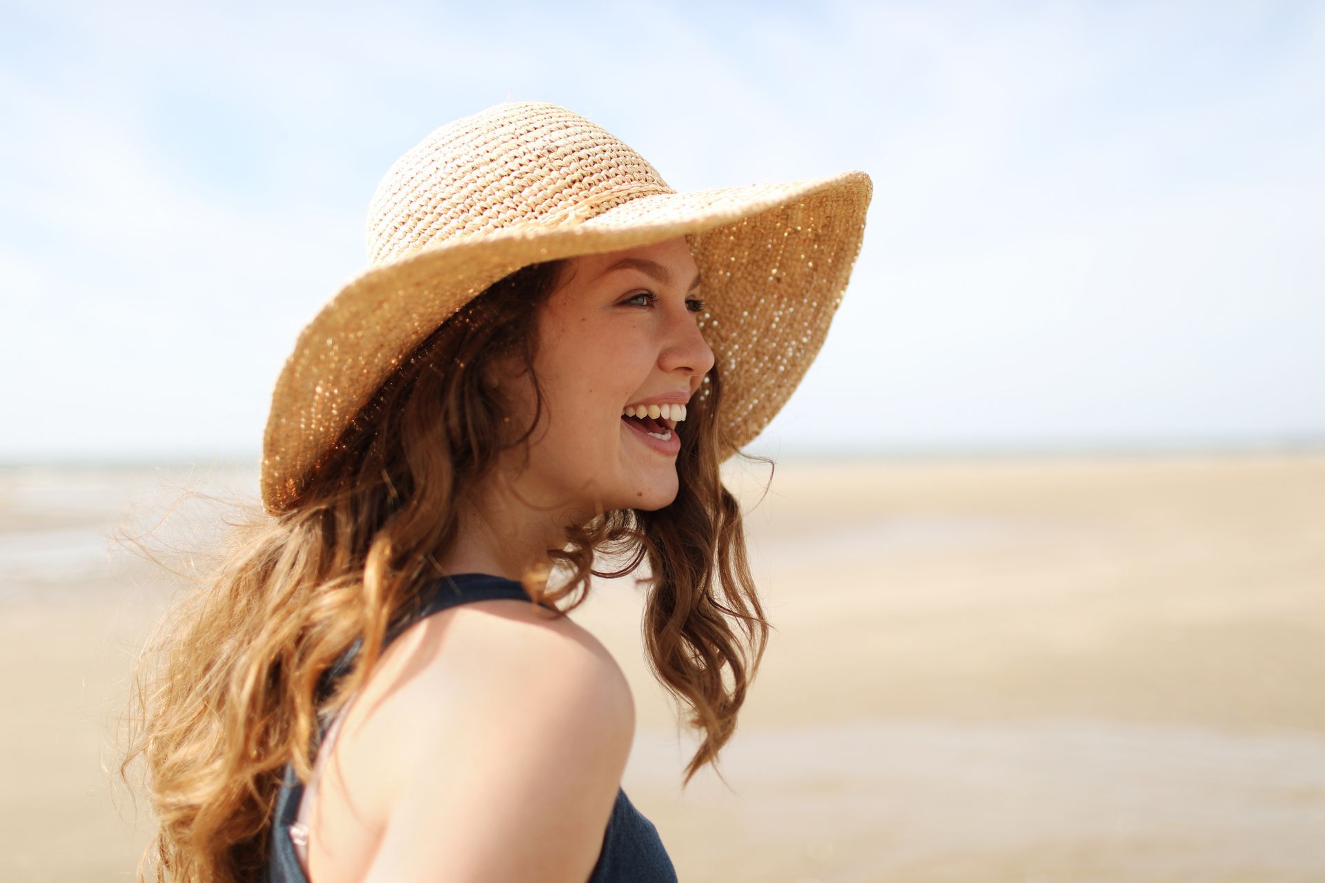 Una donna che indossa un cappello di paglia ride sulla spiaggia.