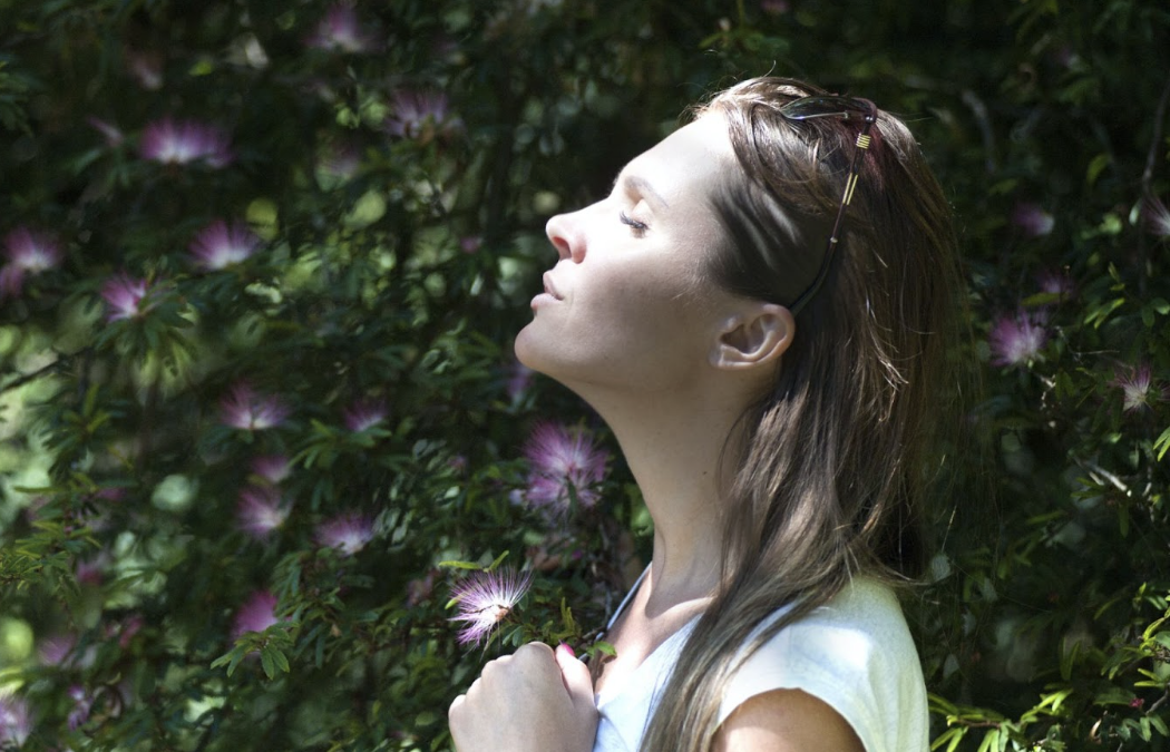 A woman is standing in front of a bush with her eyes closed.