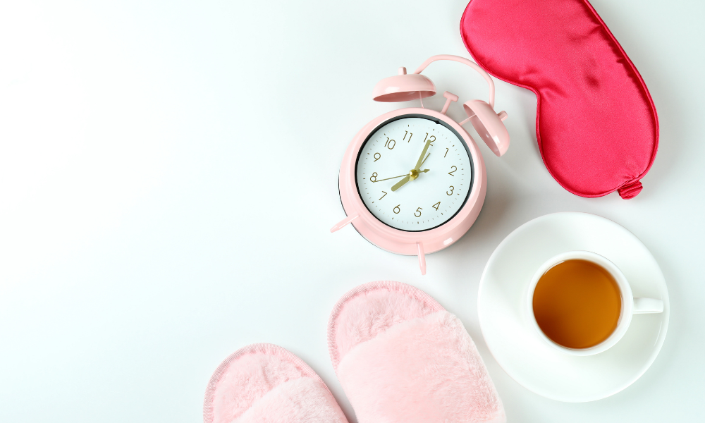 A pink alarm clock , sleep mask , slippers and a cup of coffee on a white table.