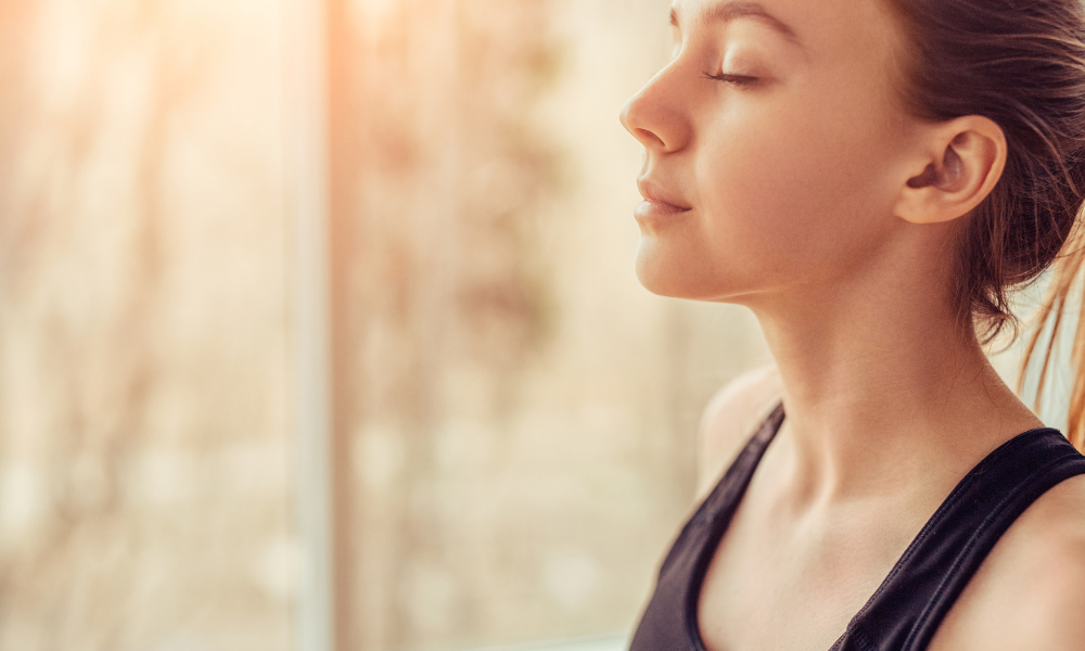 A woman is meditating with her eyes closed in front of a window.