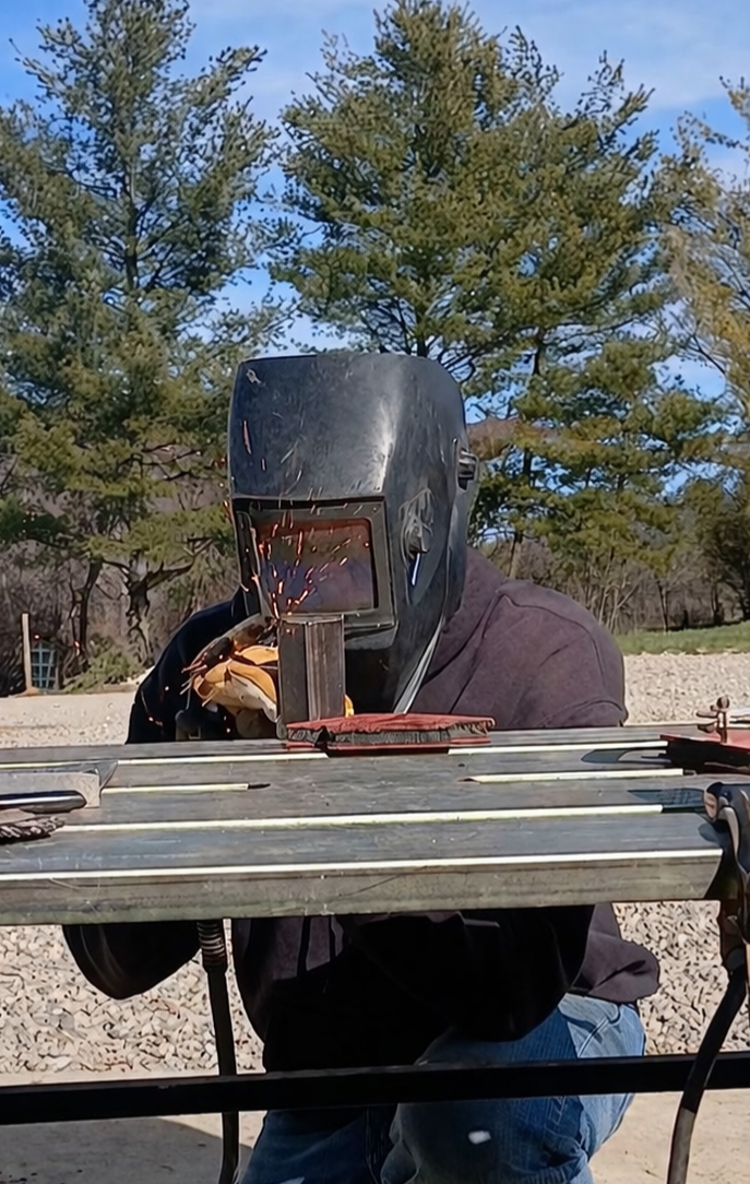 A close-up of a gloved hand using a welding torch to join metal pieces, with bright orange sparks flying in a workshop.