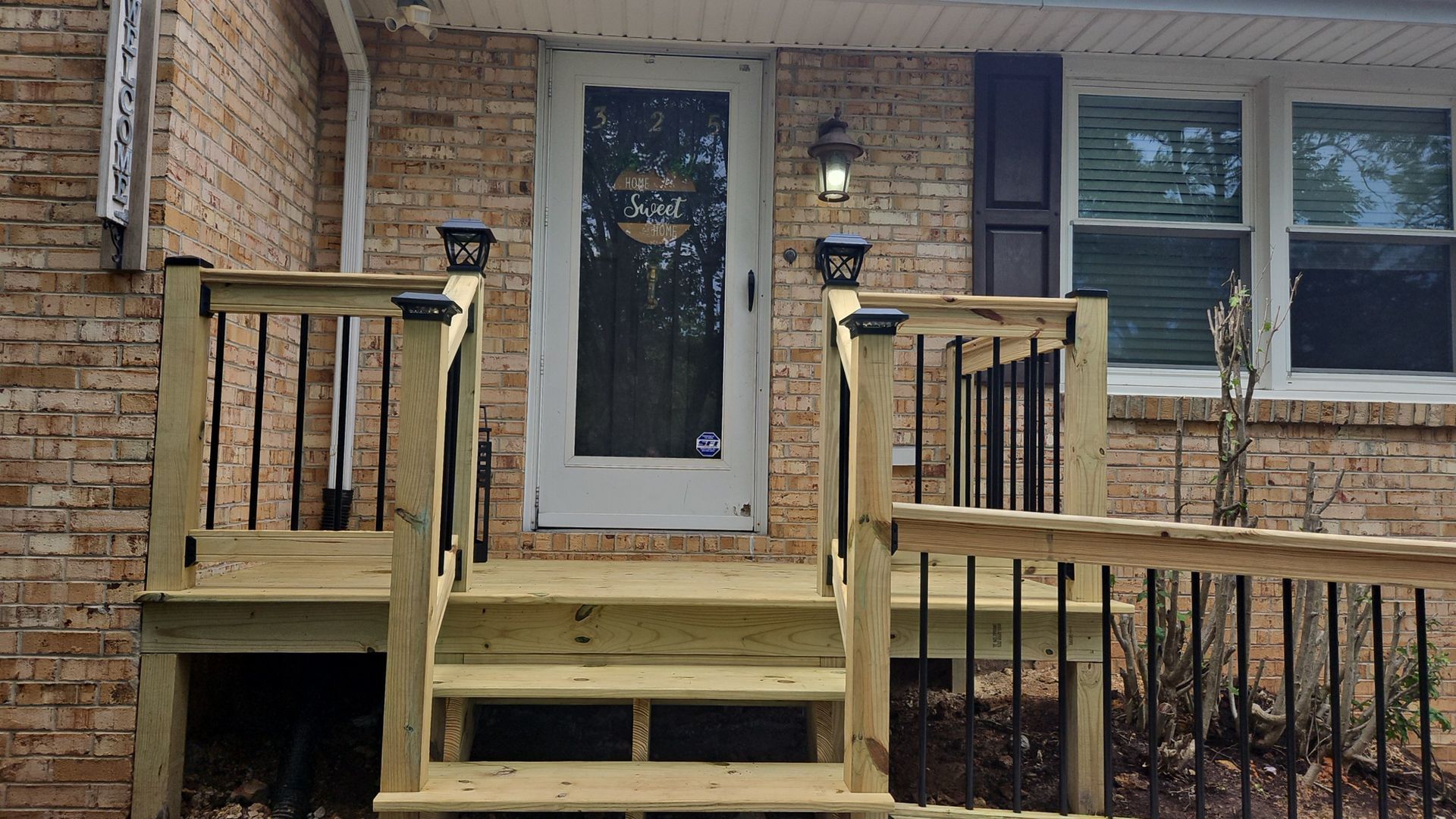 A small wooden table and two black folding chairs on a wooden deck under a pergola with string lights, overlooking trees.