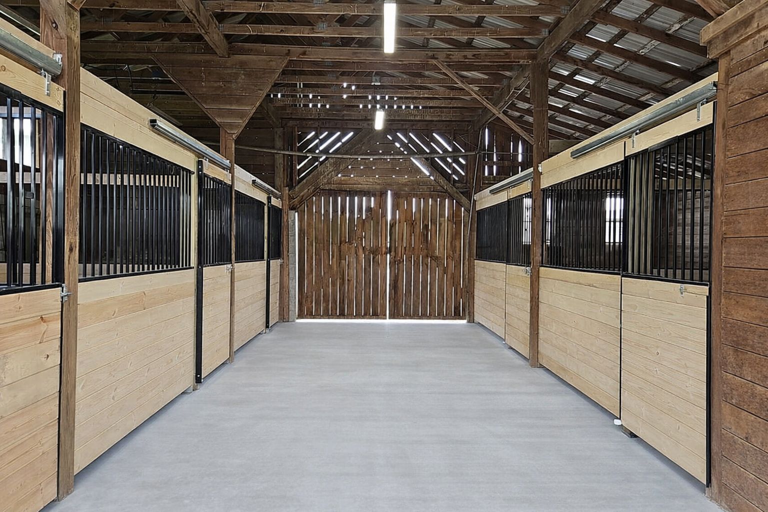 A view down the center aisle of a rustic wooden horse barn with stalls on both sides and concrete flooring.