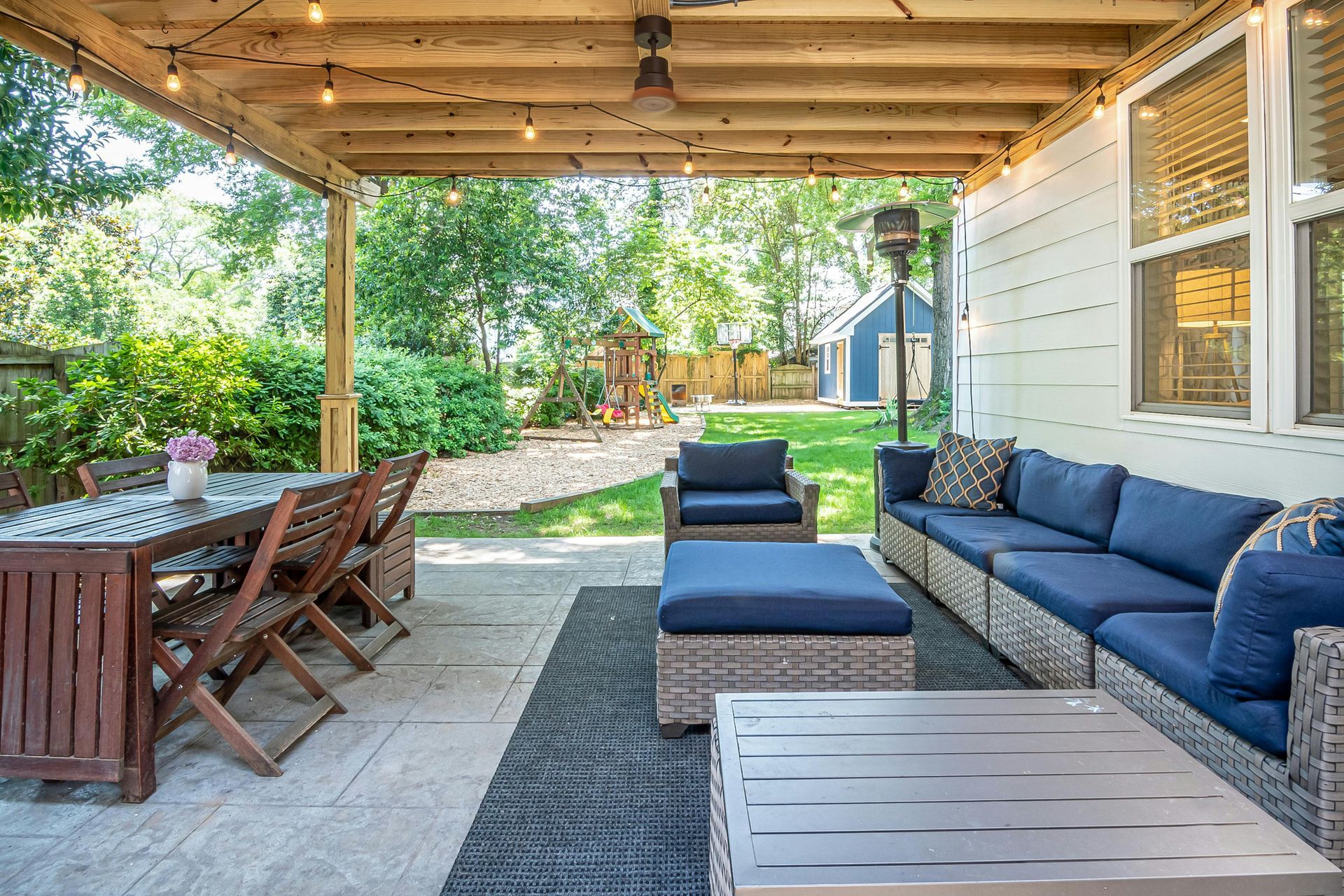 A small wooden table and two black folding chairs on a wooden deck under a pergola with string lights, overlooking trees.