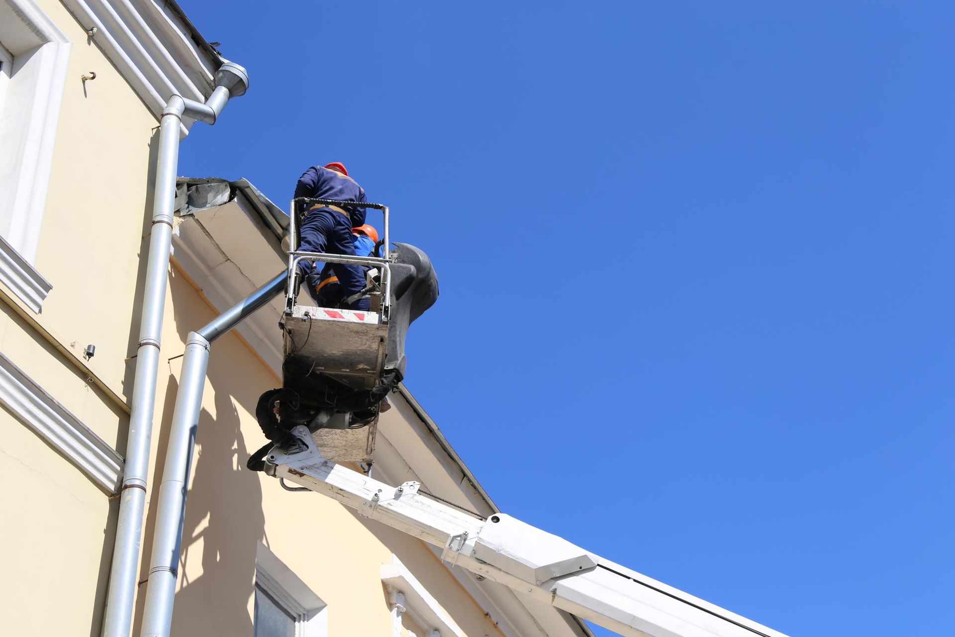 Man in a lift bucket repairing roof of a light yellow building against a bright blue sky.