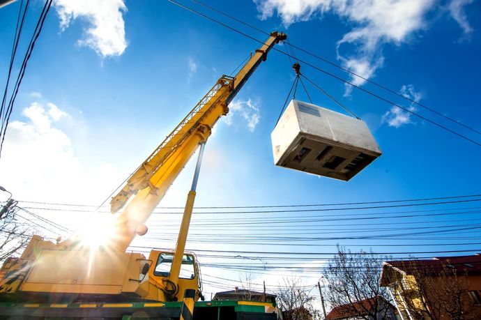 A crane lifts a white box under a bright blue sky with power lines overhead.