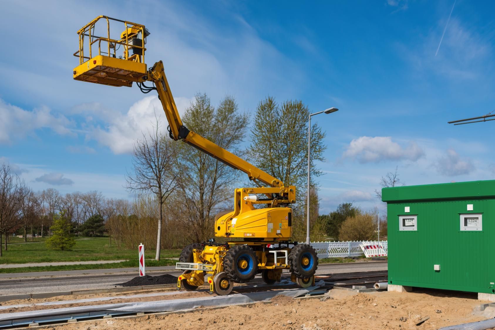 Yellow boom lift on tracks, extended toward the sky, beside a green utility building under a blue sky.