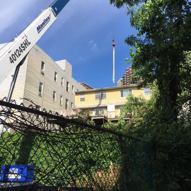 A crane lifting a long white object over a yellow building, behind a chain link fence.